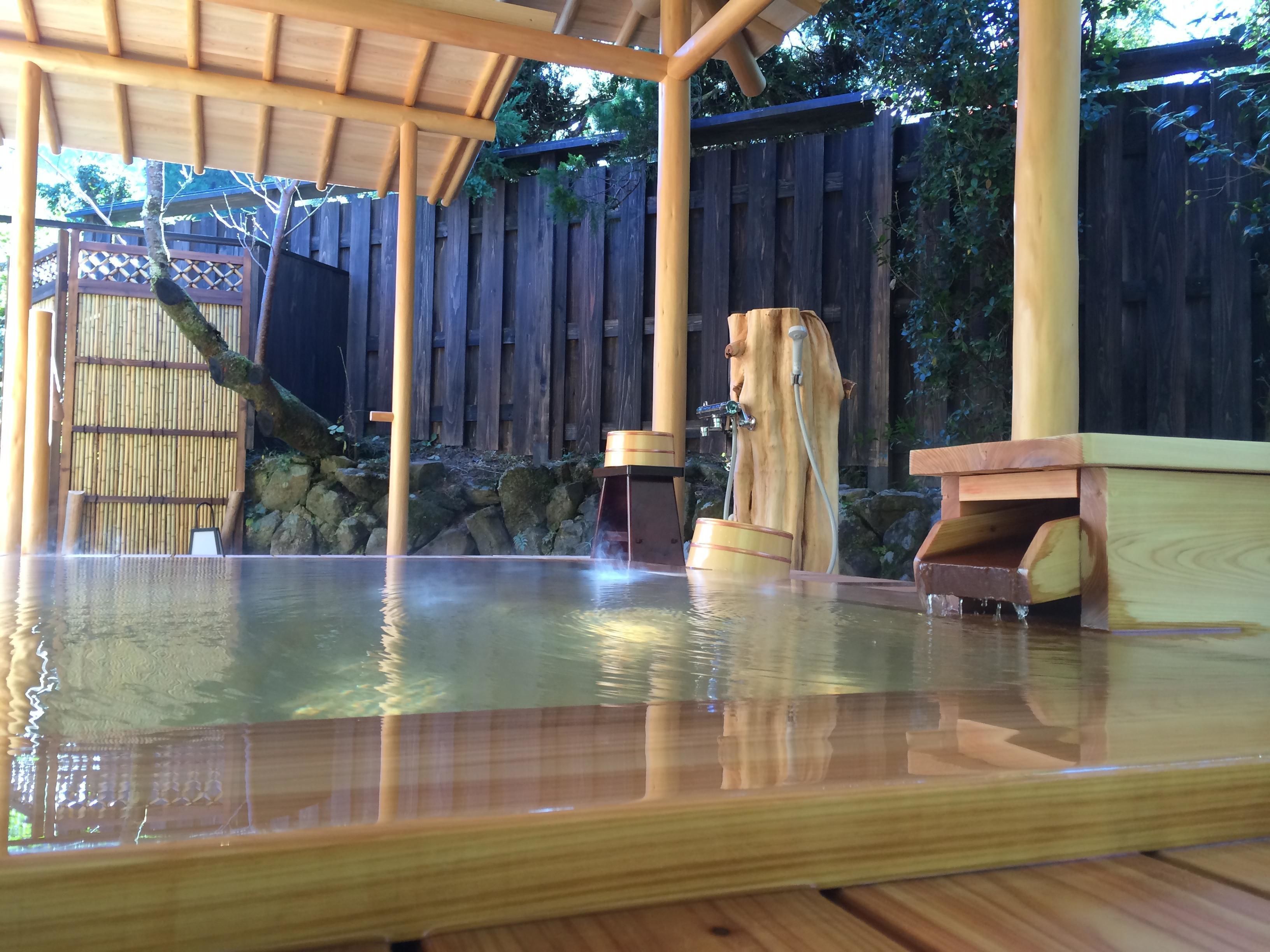 A wooden hot spring bath area with a wooden structure overhead, a bamboo fence, and a wooden deck.