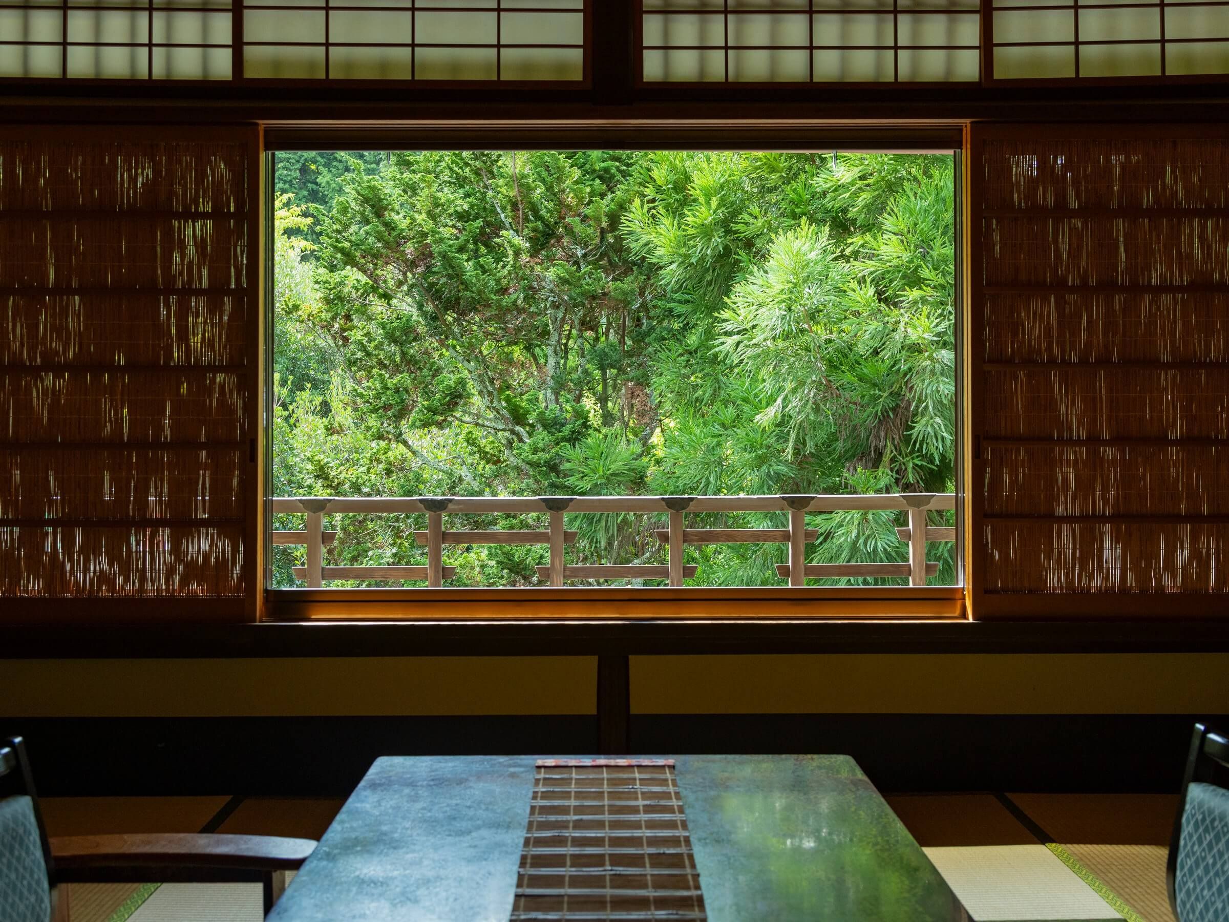 A room with tatami flooring, a low table, and a large window with a view of dense green foliage.