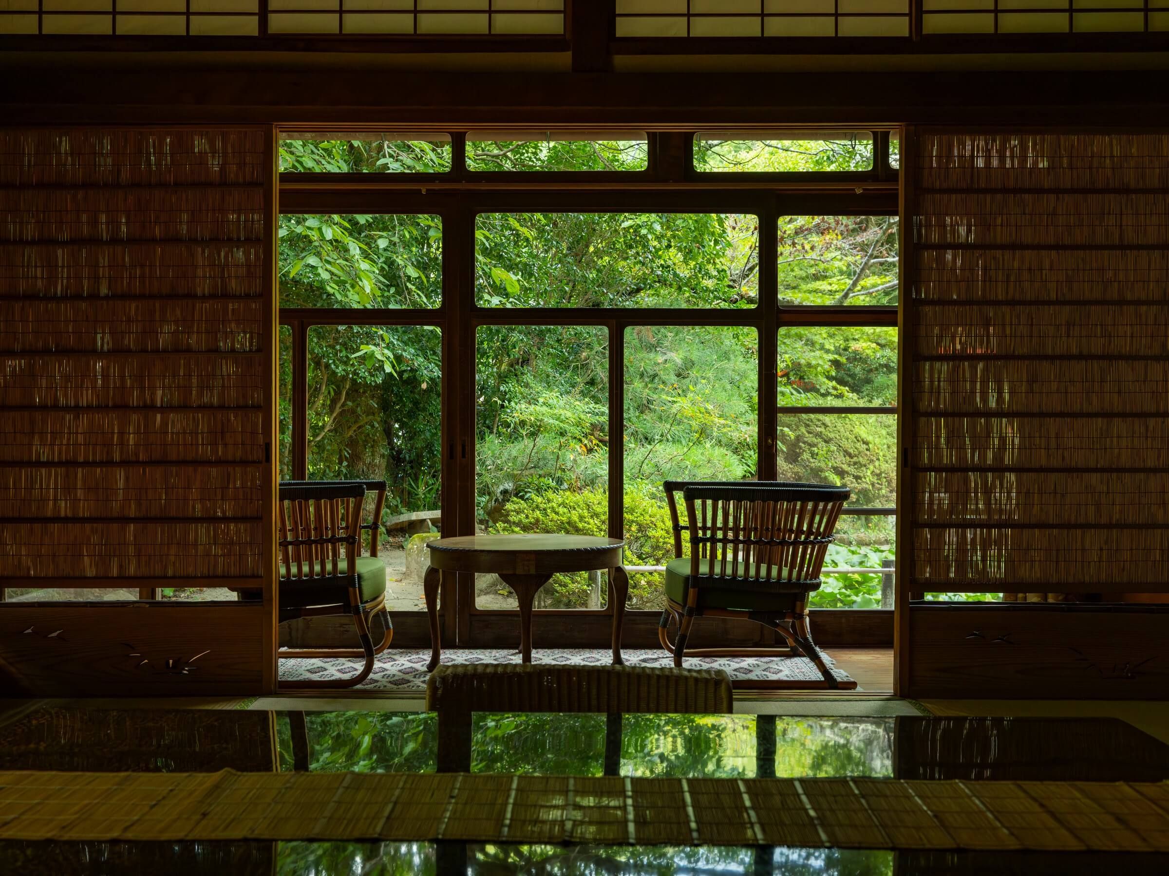 A room with a view of a lush garden through large windows, a table, and two chairs.