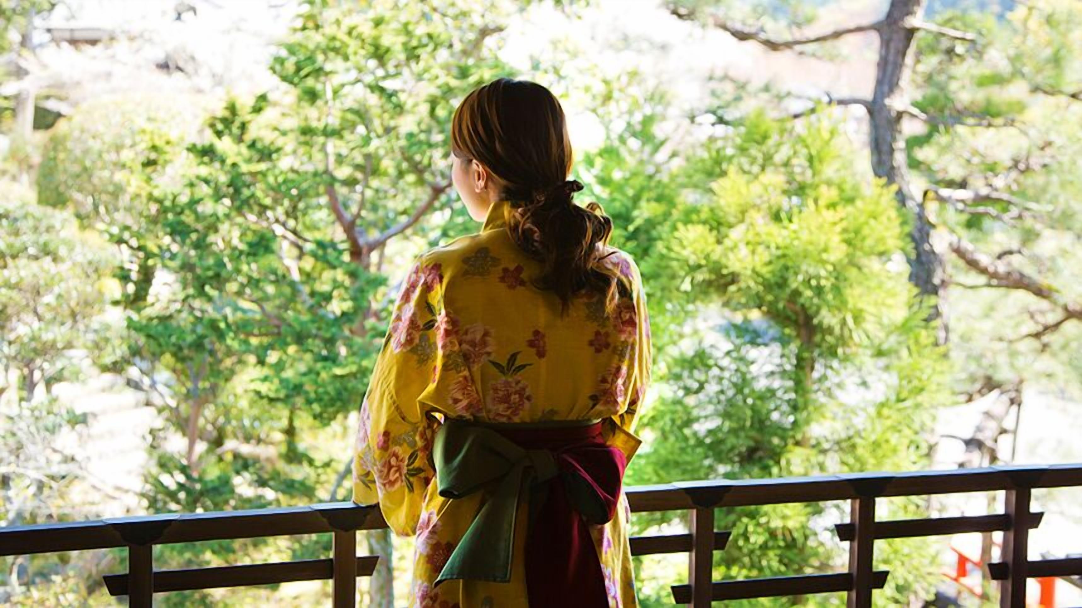 A person in traditional attire standing on a balcony with a view of trees.
