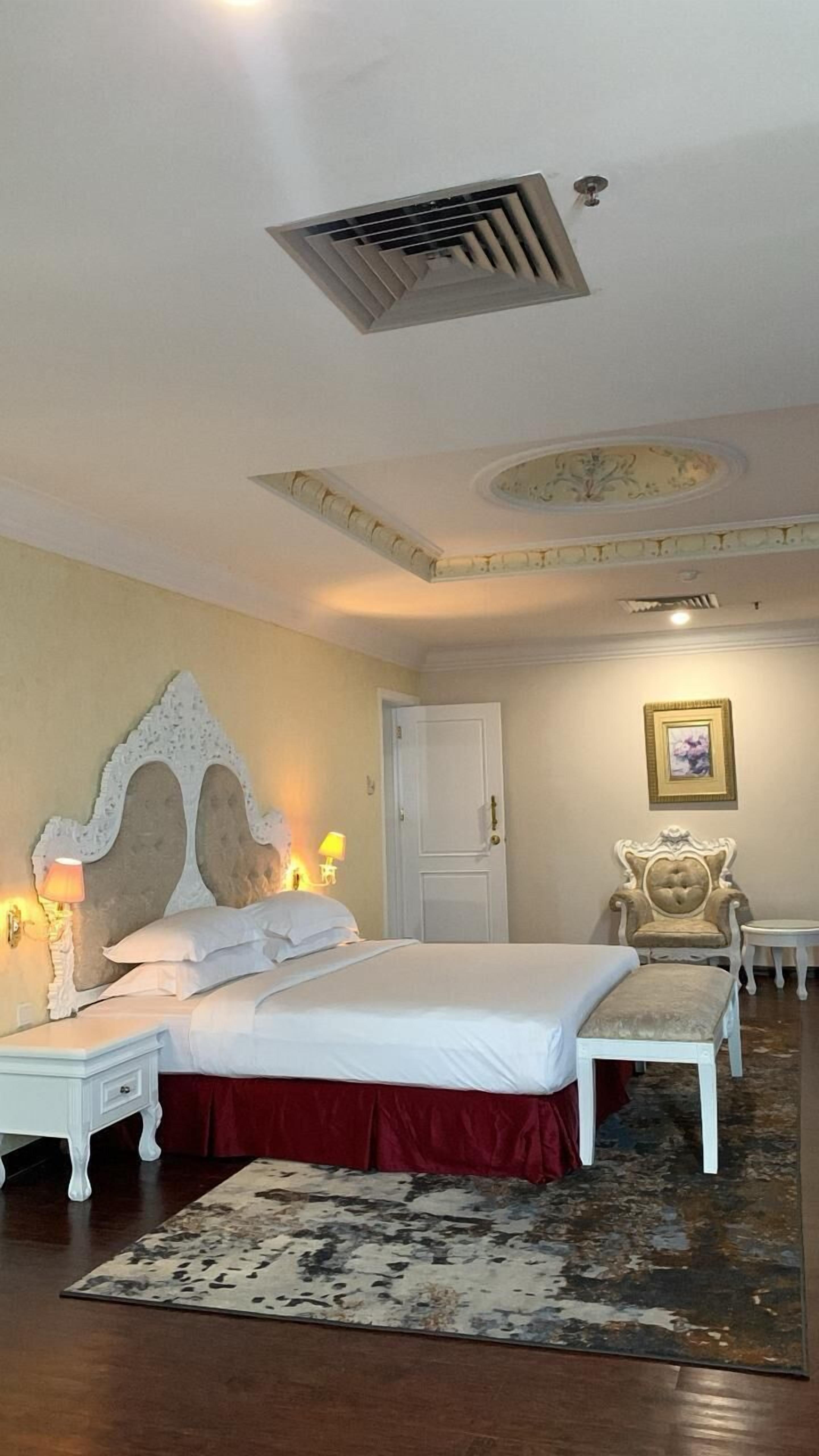 Une chambre d’hôtel avec un grand lit, une table de chevet, une chaise, un tapis et une bouche d’aération au plafond.