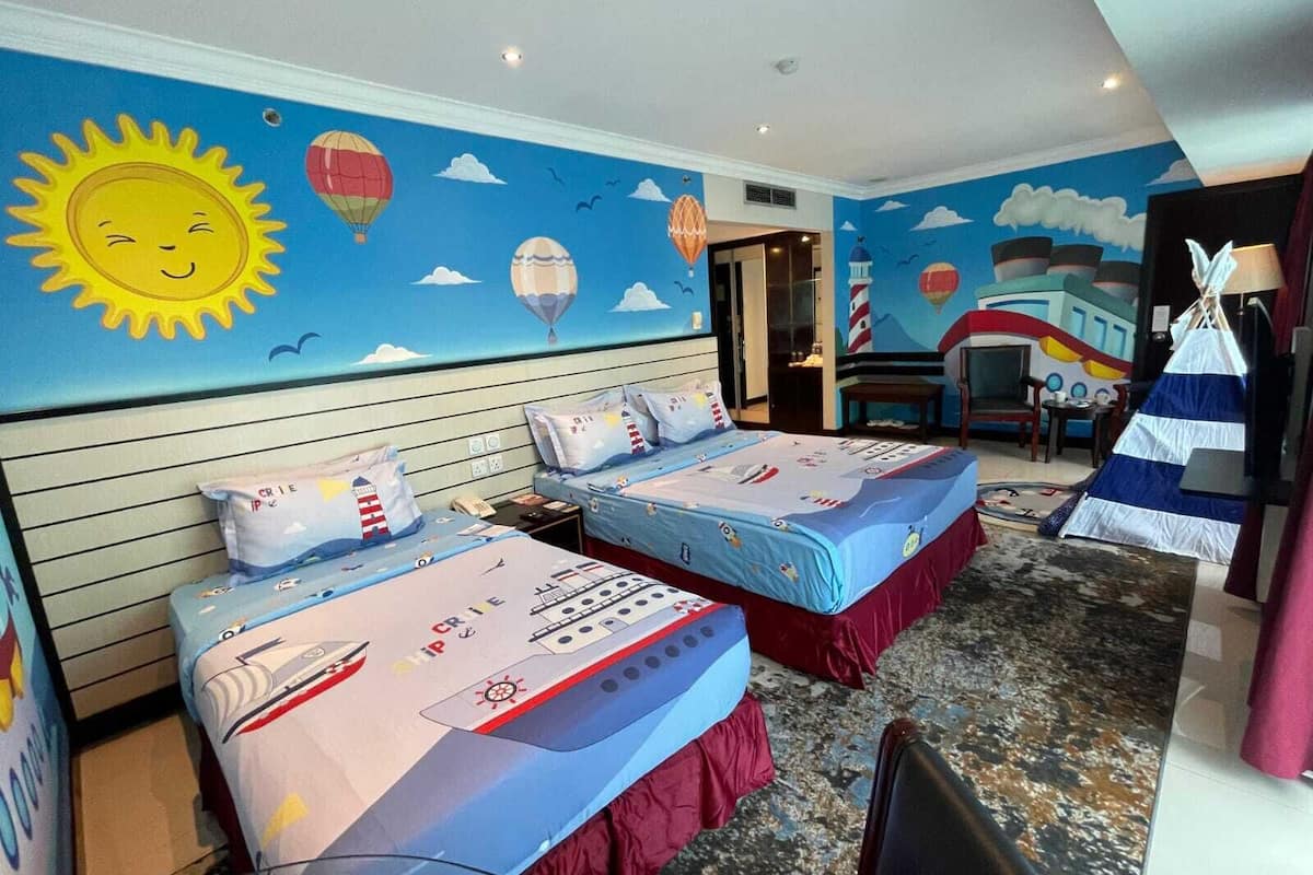 A hotel room with two beds, nautical-themed bedding, a mural of the sky with clouds and hot air balloons, and a small table with a lamp.
