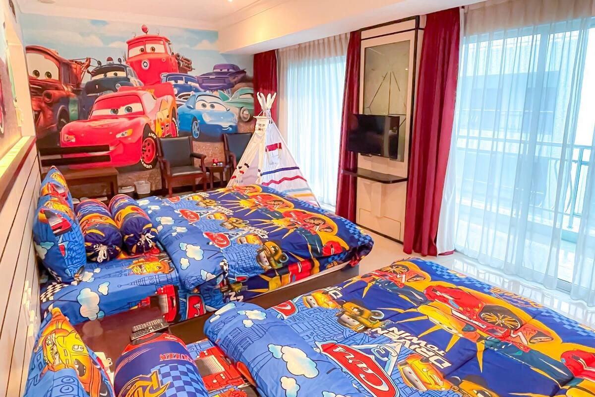 A hotel room with a bed featuring a Cars-themed duvet cover, a large mural of Cars characters on the wall, a TV mounted on the wall, and a window with curtains.