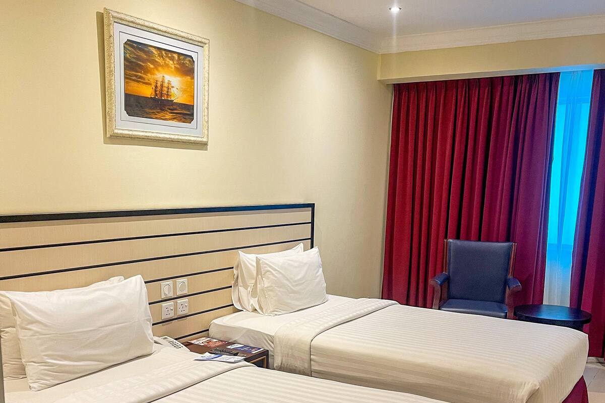 A hotel room with two beds, a desk, a chair, and a window with red curtains.