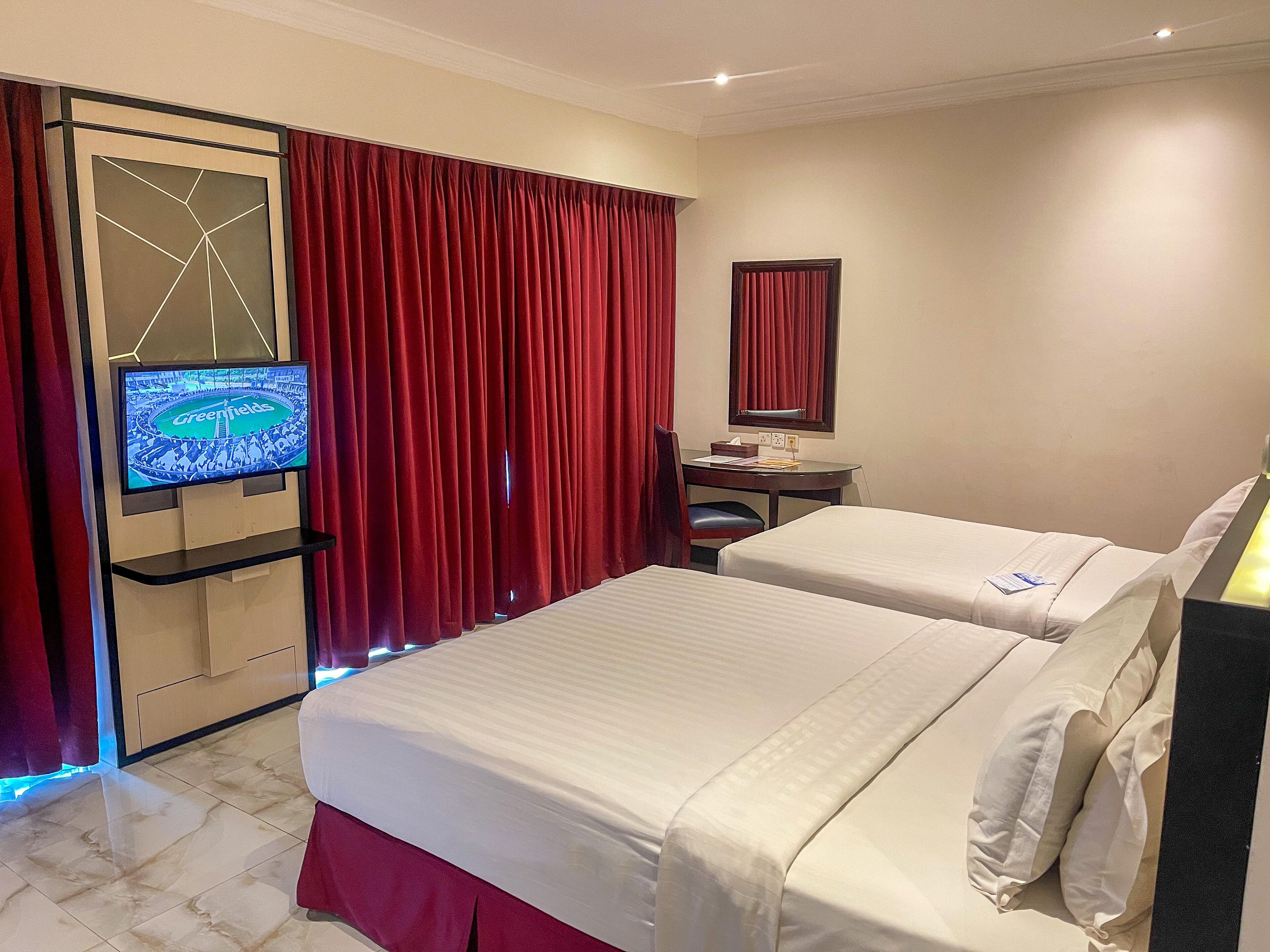 Habitación de hotel con dos camas, un televisor en un mueble bajo, un escritorio con una silla y cortinas rojas.