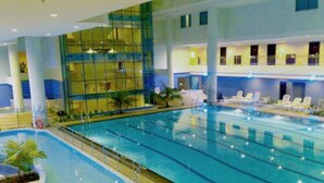 A large indoor swimming pool with multiple lanes, surrounded by lounge chairs and a glass-enclosed area with a view of the interior. - Pacific Palace Hotel (Batam)