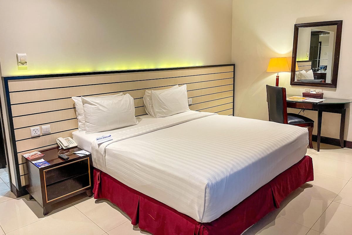 A hotel room with a large bed, a bedside table with a phone and magazines, a desk with a lamp, and a mirror on the wall.