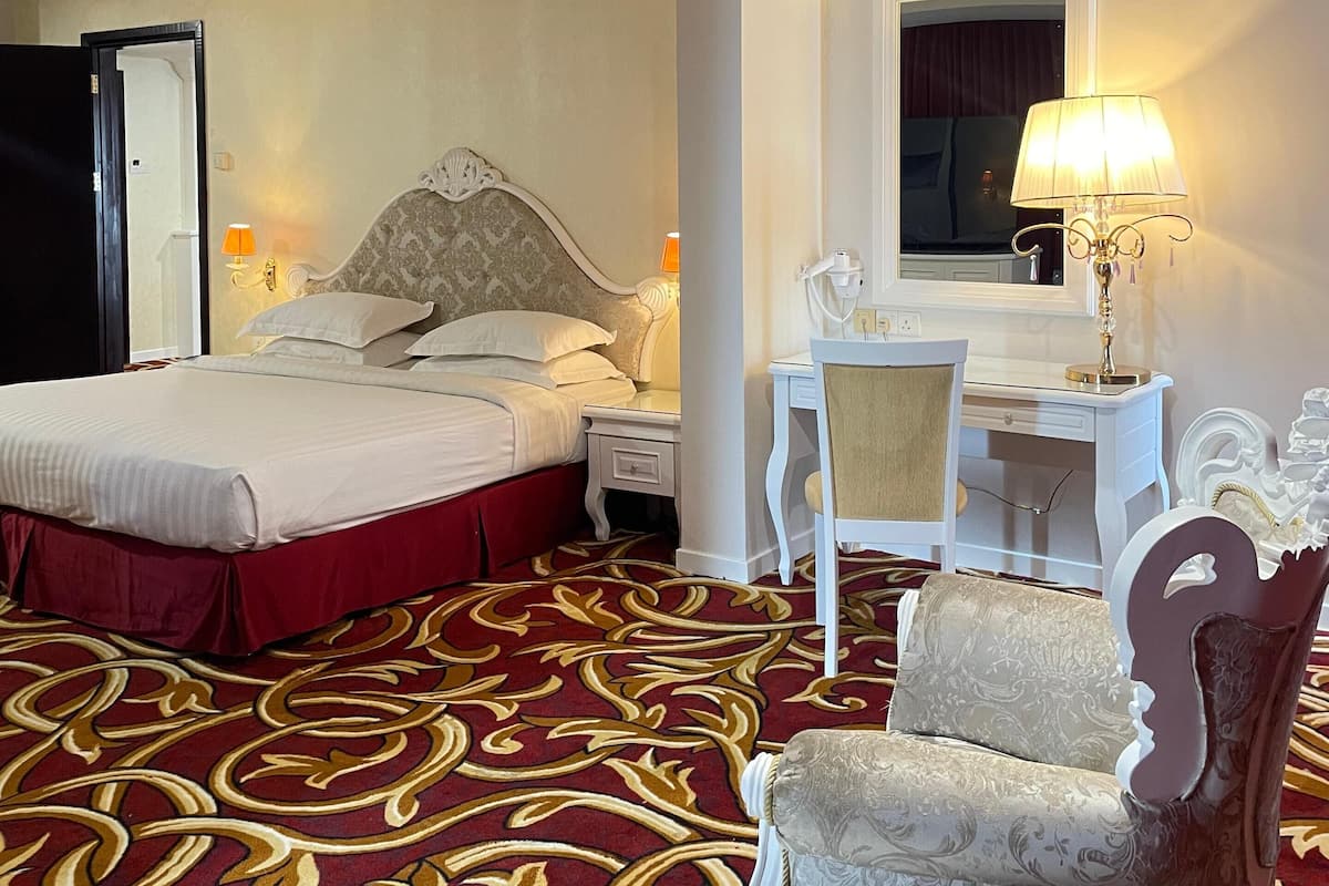 A hotel room with a large bed, a desk with a chair, a desk lamp, and a decorative chair.