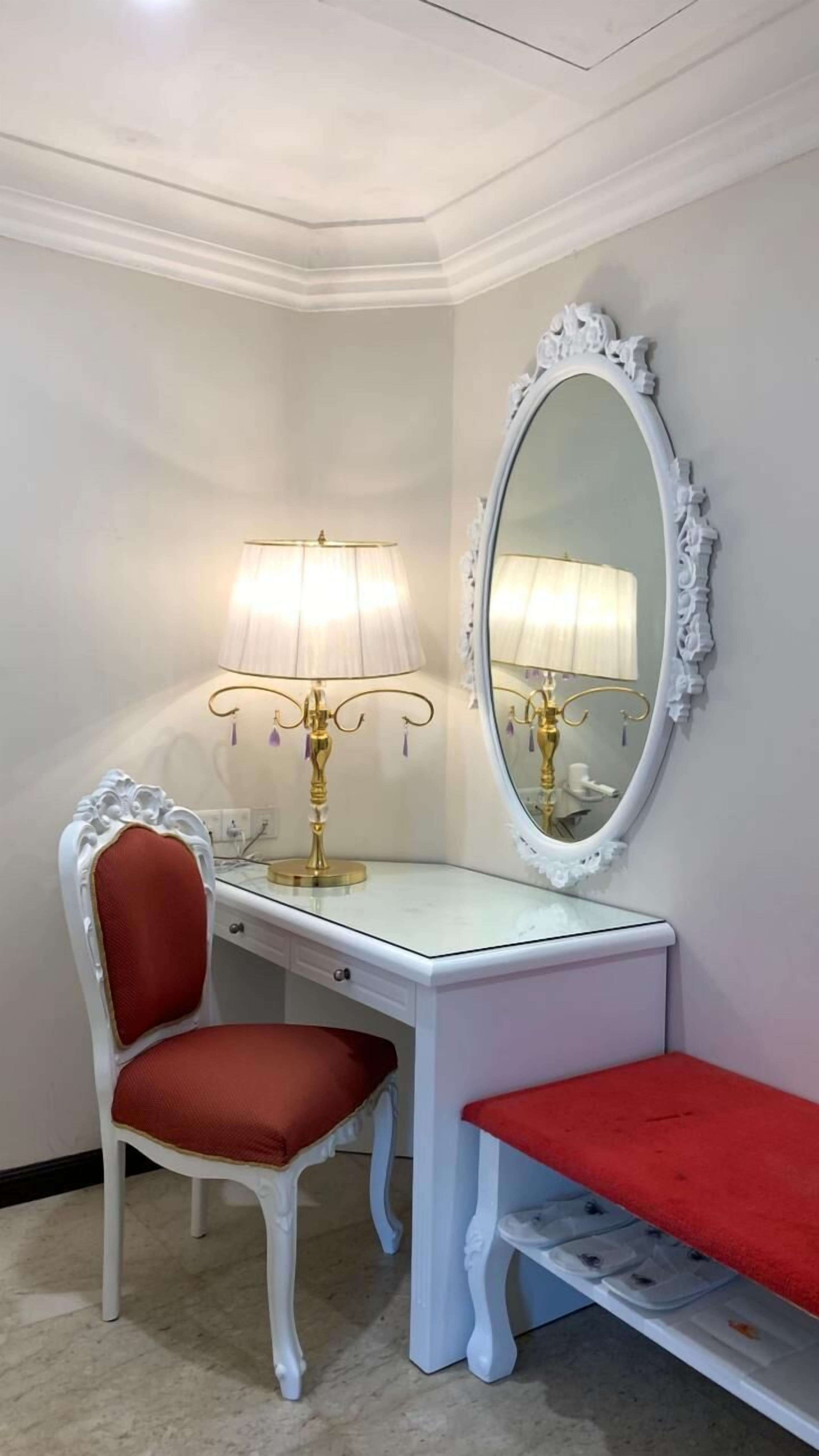 A white vanity with a red cushioned chair and bench, a gold lamp, and a large ornate mirror.