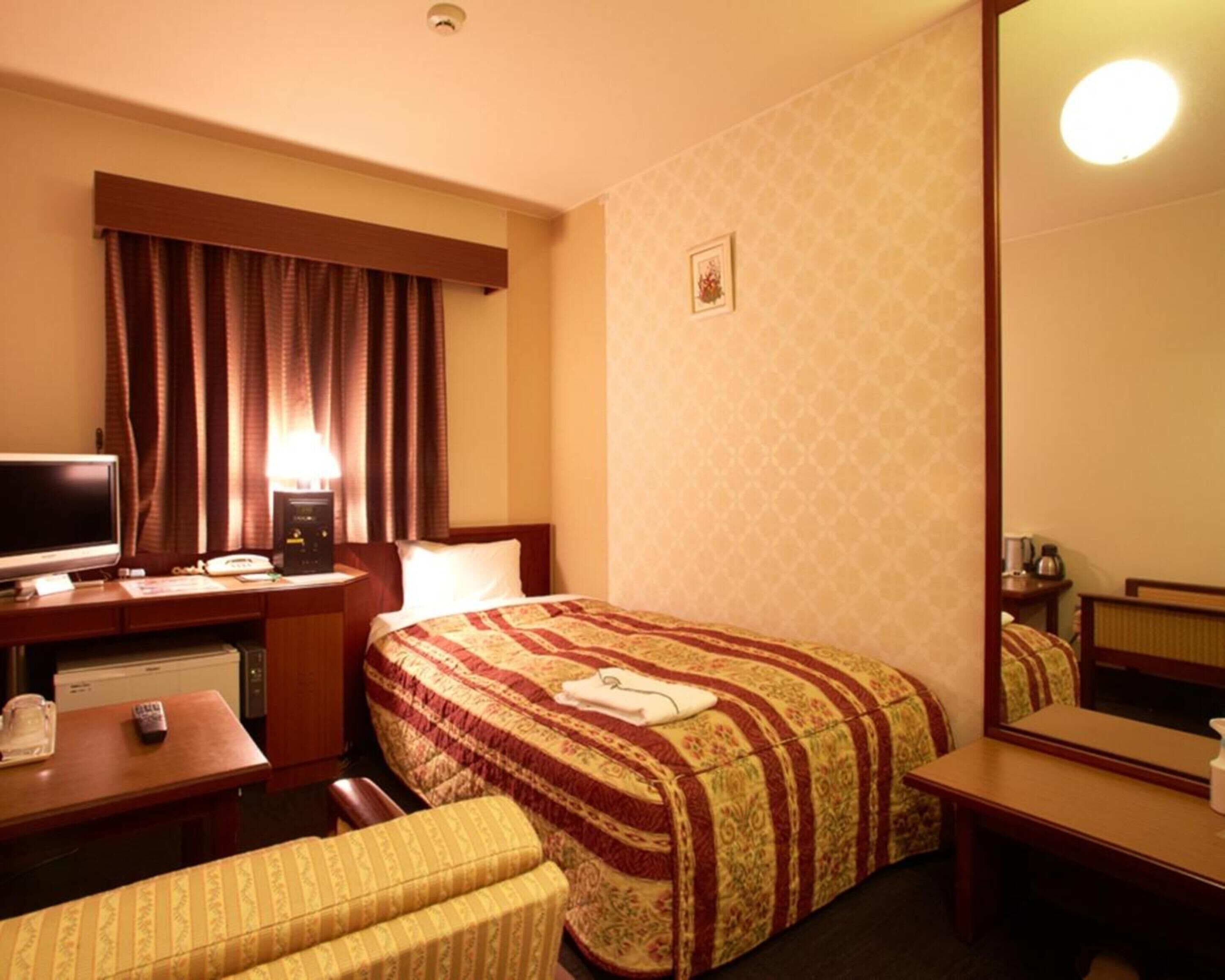 Habitación de hotel con cama, escritorio con computadora, silla, mesita de noche, lámpara y espejo.