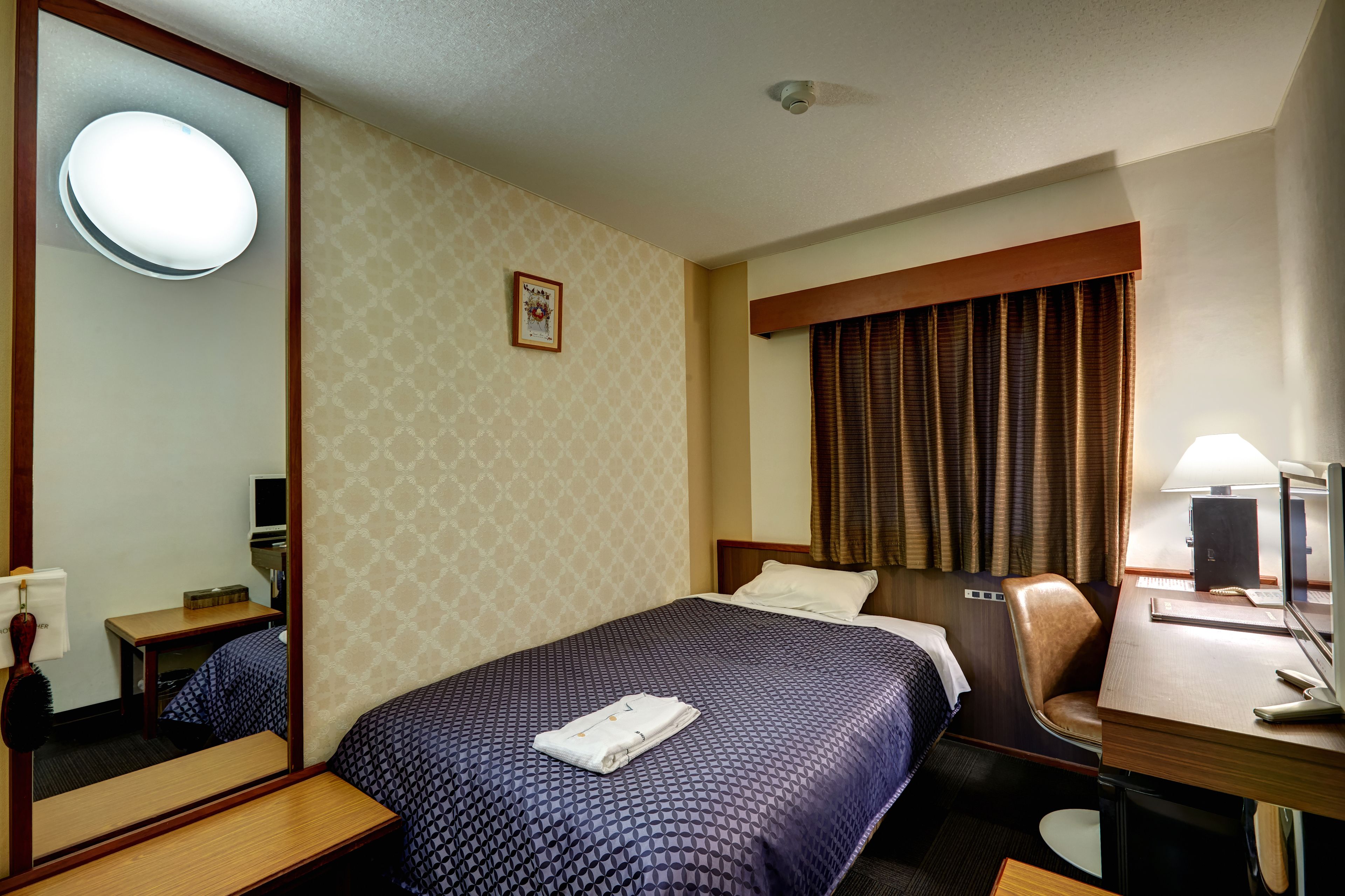 Habitación de hotel con una cama grande, un escritorio con computadora, una silla, un espejo y una ventana con cortinas.