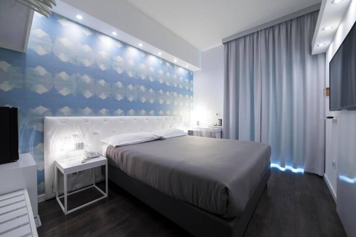 A modern hotel room with a large bed, a bedside table with a lamp, a wall-mounted TV, and a large window with curtains.
