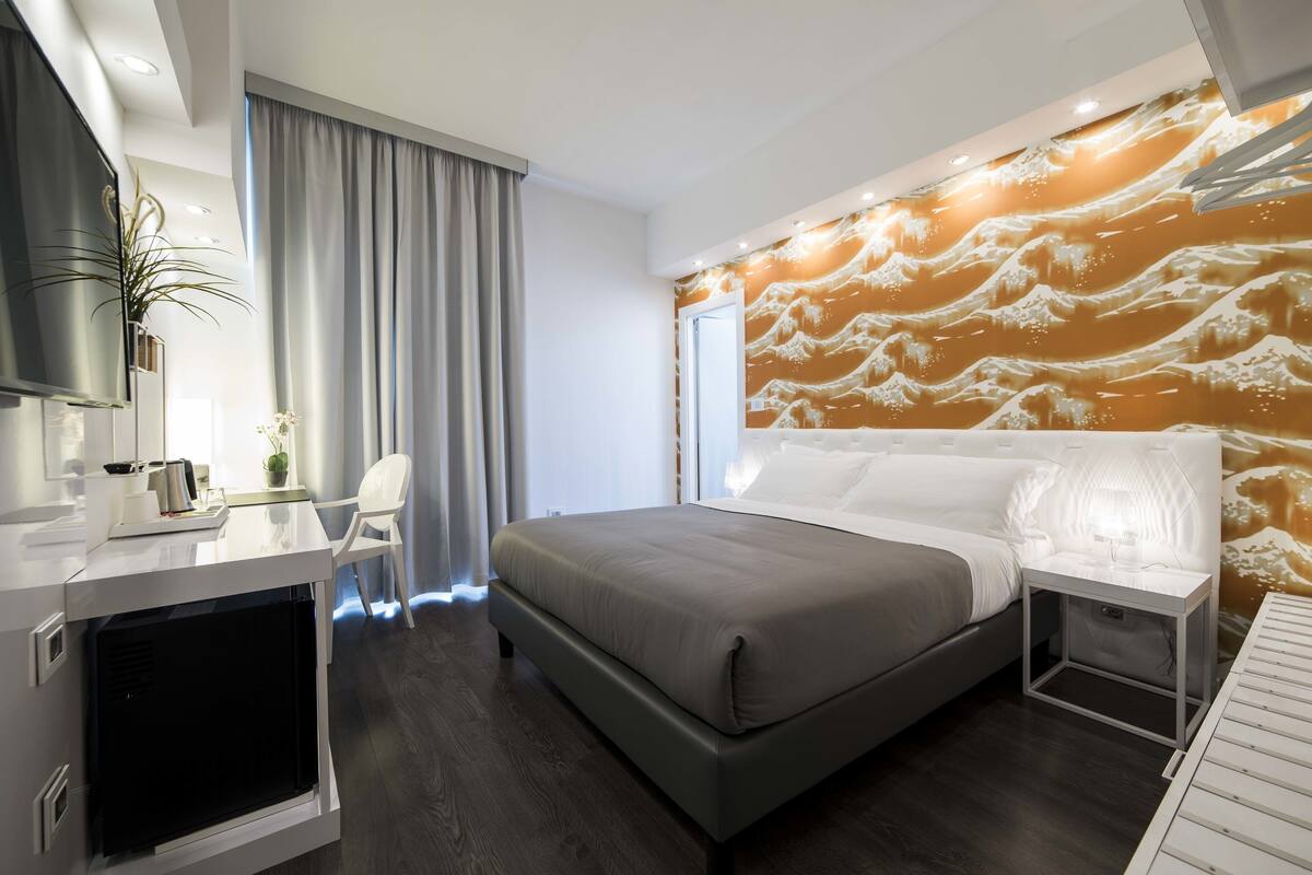 A modern hotel room with a large bed, a desk with a flat-screen TV, and a wall with a wave pattern design.