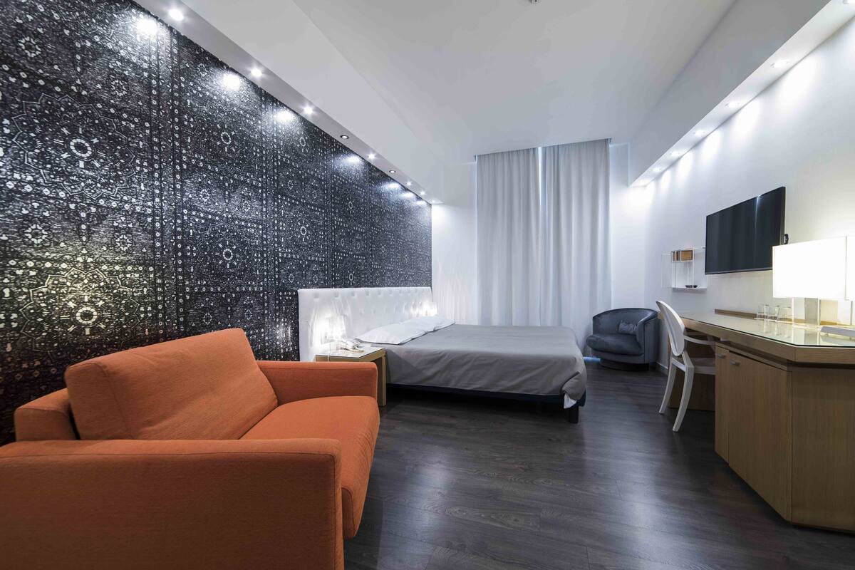 A modern hotel room with a bed, a desk, a chair, and a sofa.