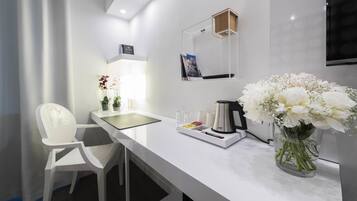 Una camera d'albergo moderna con una scrivania bianca, una sedia, una macchina per il caffè, un vaso con fiori e una TV a schermo piatto montata a parete.