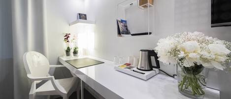 Quarto de hotel moderno com escrivaninha branca, uma cadeira, cafeteira, um vaso com flores e uma TV de tela plana fixada na parede.