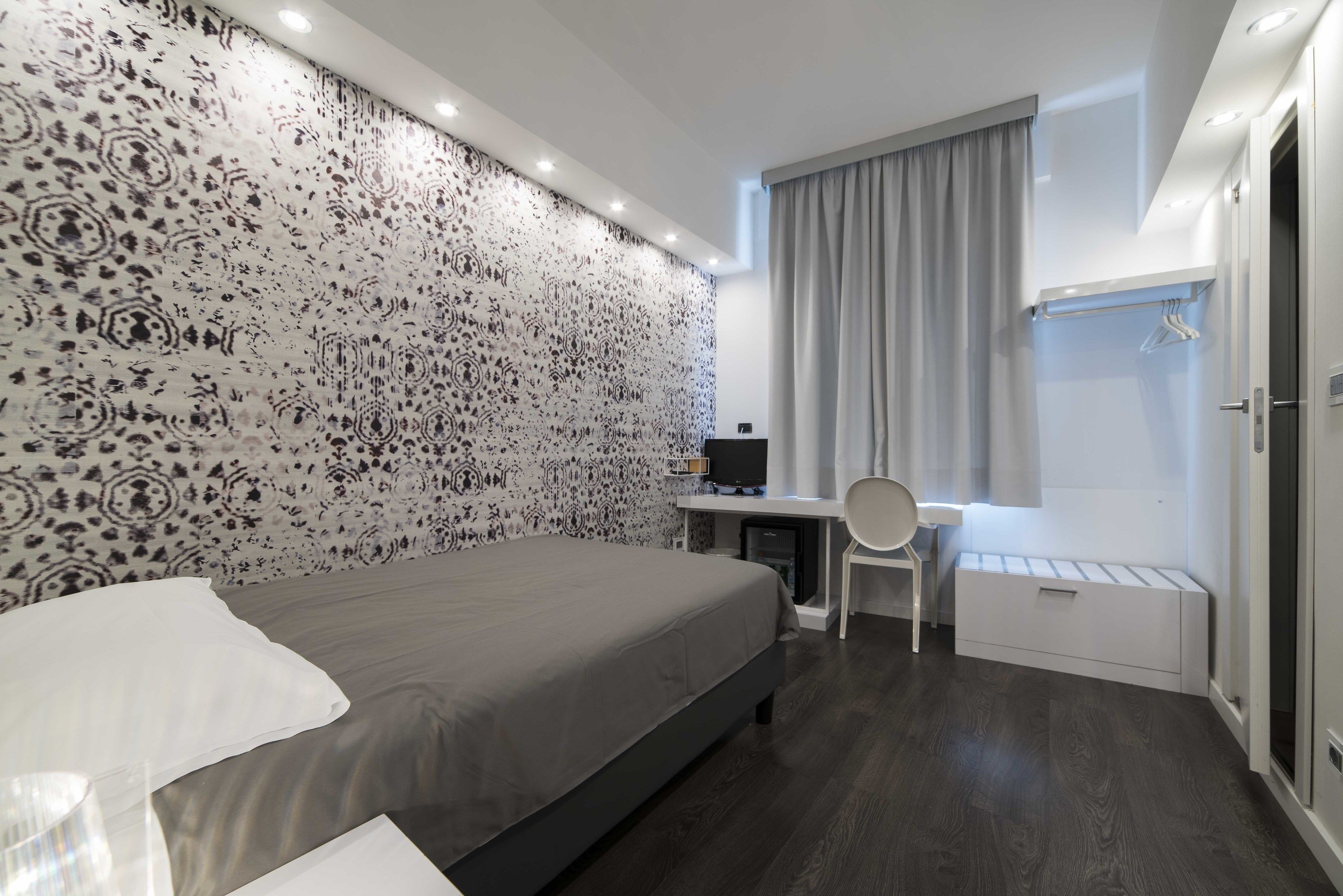 A modern hotel room with a large bed, a desk with a chair, a small table, and a wardrobe.