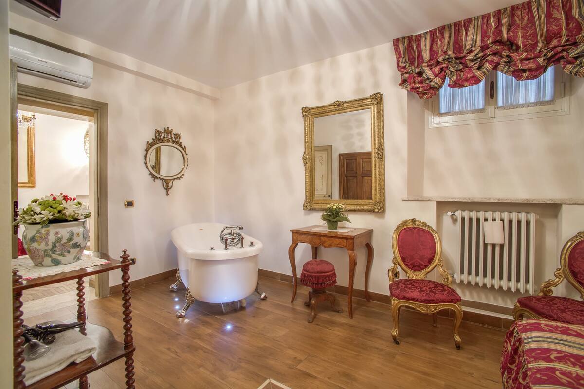 A classic bathroom with a freestanding bathtub, a vanity with a mirror, and two ornate chairs.