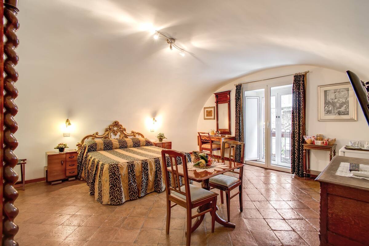 A bedroom with a bed, a dining table with chairs, a small table with a lamp, and a large window with curtains.