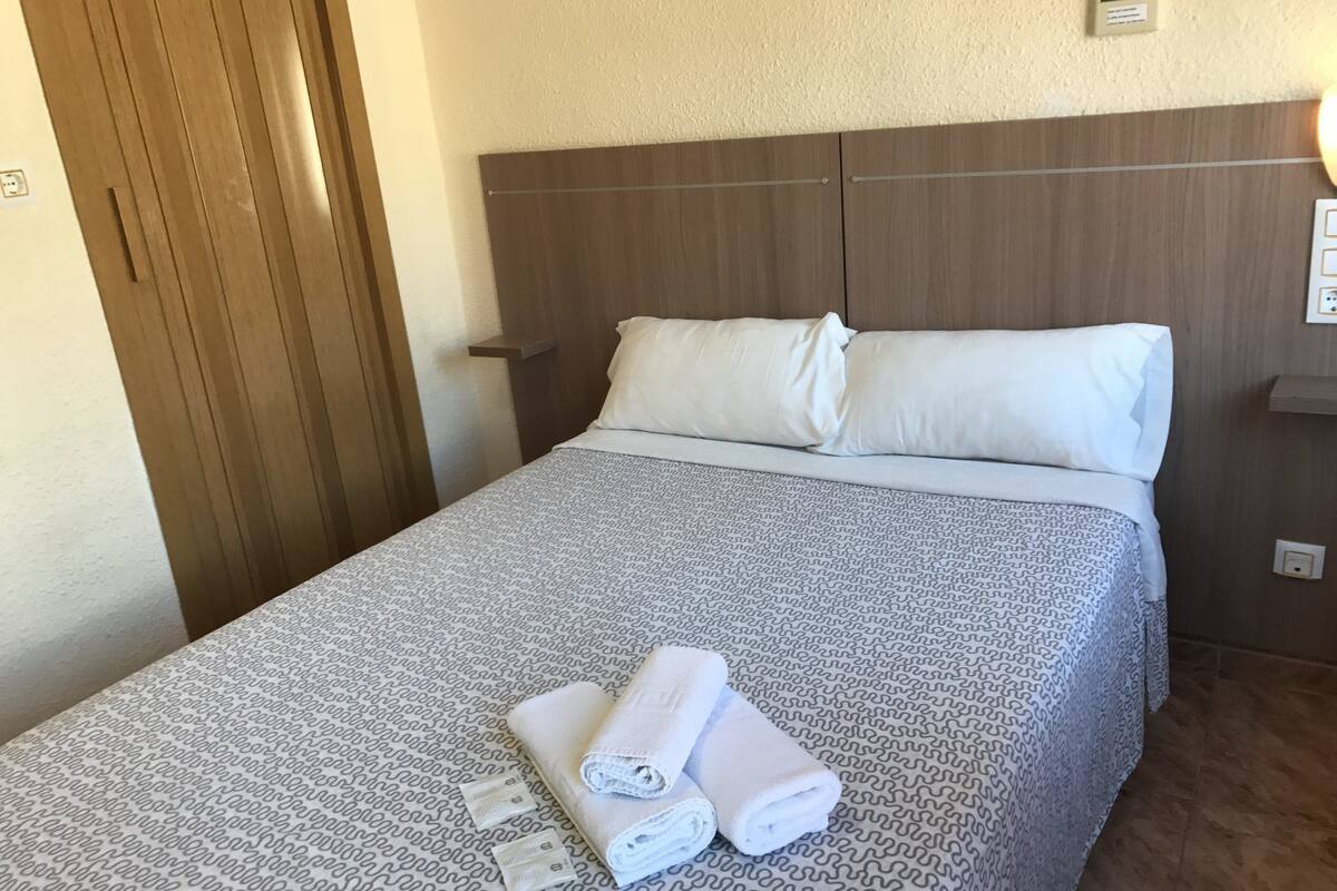 A neatly made bed with a patterned bedspread, two pillows, and two rolled towels on the bed.