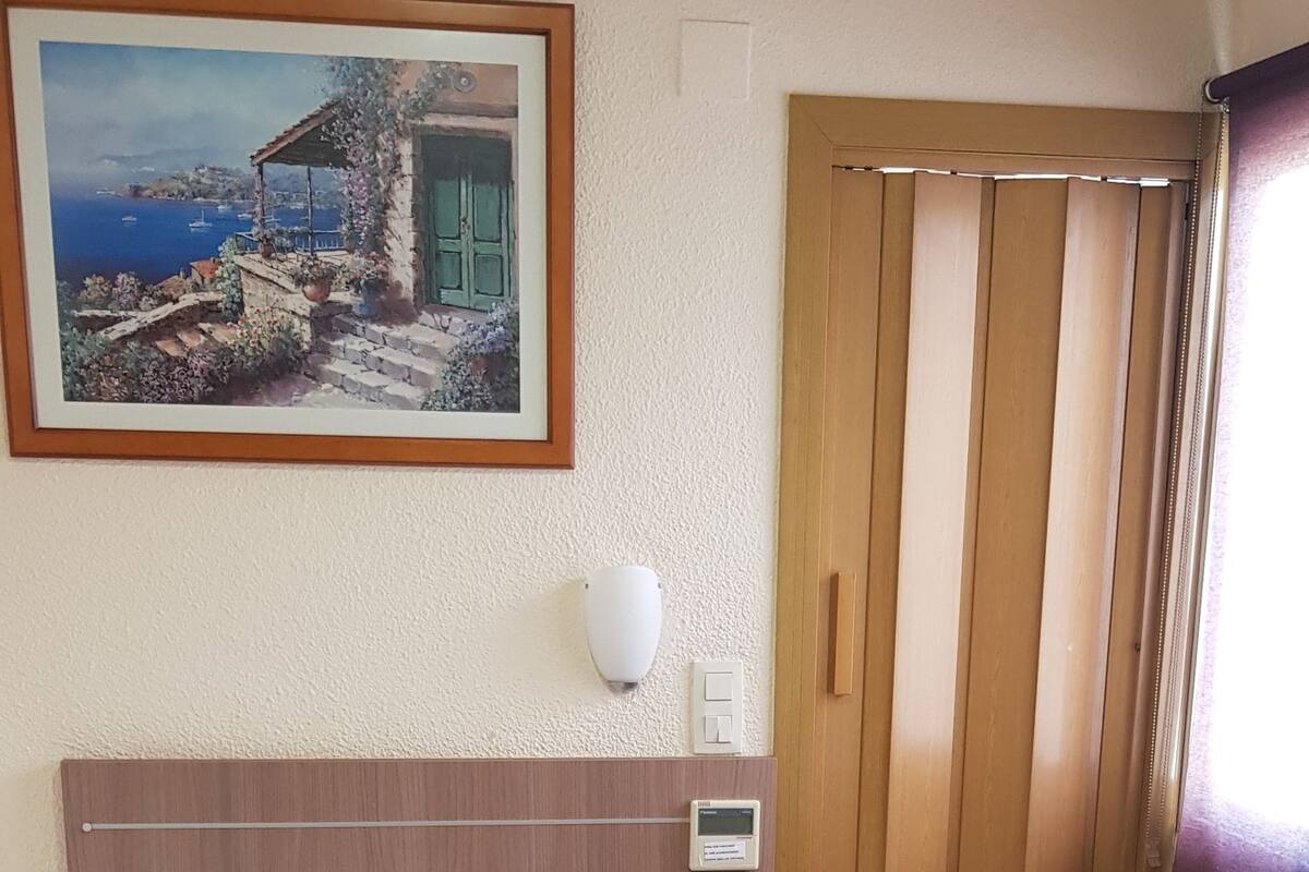 A hotel room with a bed, bedside table, lamp, and a painting of a coastal scene.