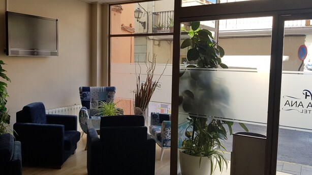 Un hall d’hôtel doté d’un téléviseur à écran plat fixé au mur, de plusieurs fauteuils bleu foncé, d’une table basse à motifs et de plantes en pot.