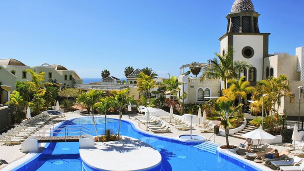 Zona de piscina en un resort con sillones de descanso, palmeras y un edificio con cúpula.