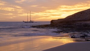 Una playa con un velero, costa rocosa y aguas tranquilas que reflejan la puesta de sol.