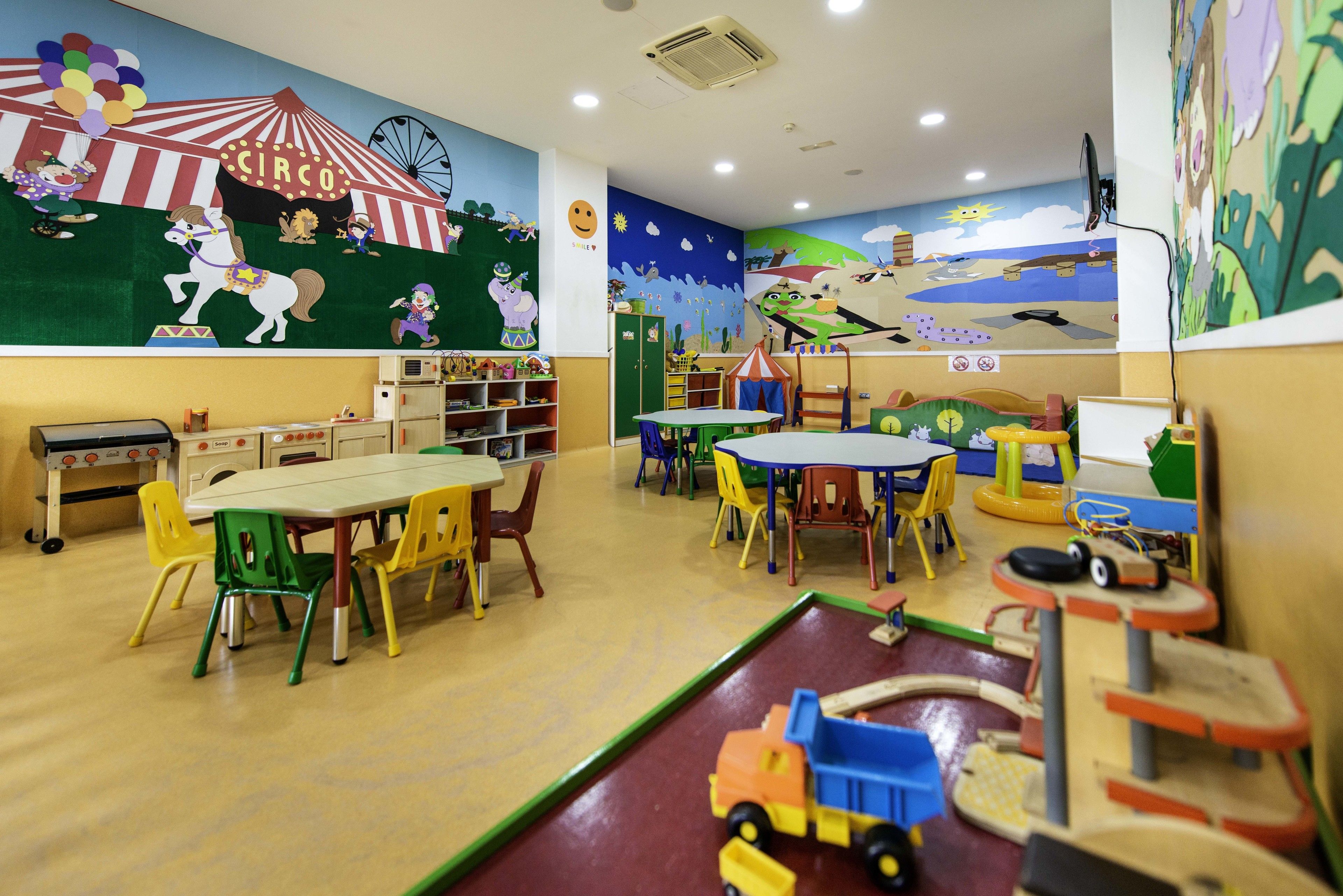 Una sala de juegos infantil con muebles coloridos, un mural con temática de circo y diferentes zonas de juegos y juguetes.