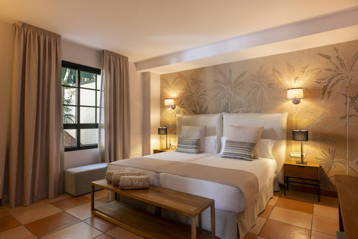 Un dormitorio amplio con una cama grande, dos mesitas de noche con lámparas, un banco y una ventana con cortinas.
