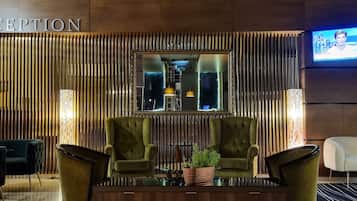 A reception area with a wooden reception desk, two green armchairs, a potted plant, and a television screen on the wall.