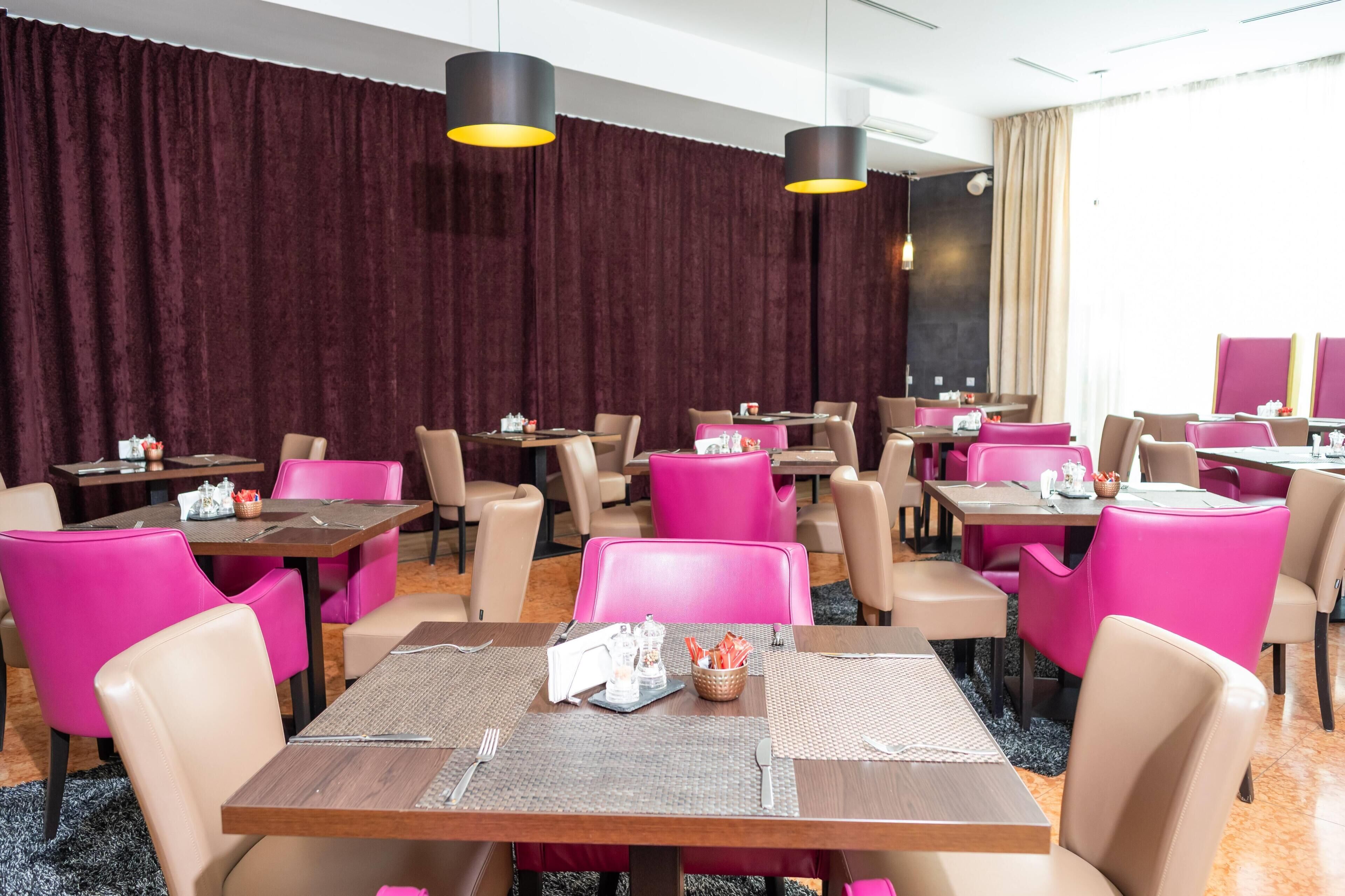 A restaurant with multiple tables and chairs, some with pink cushions, and black pendant lights.