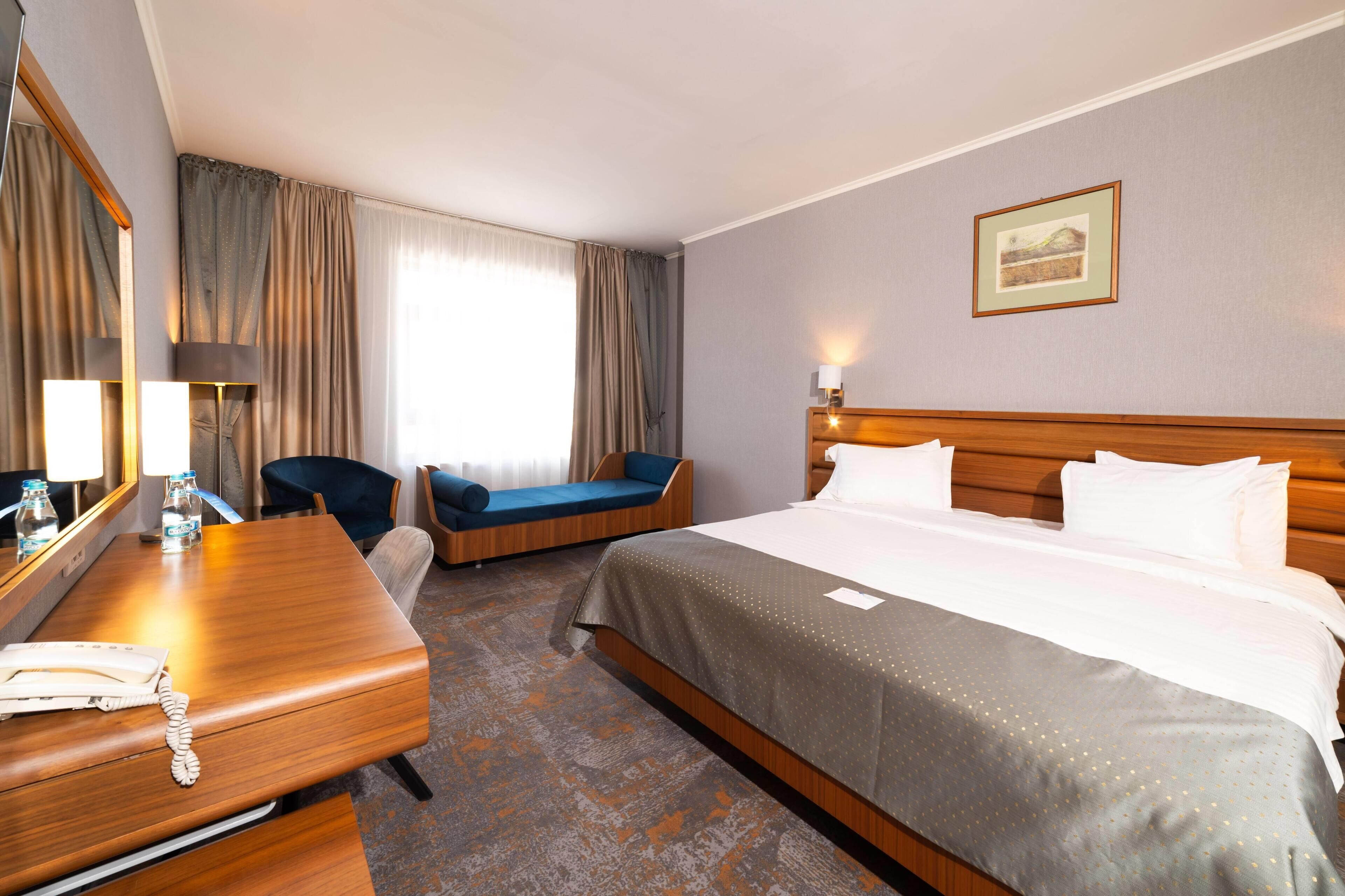 A hotel room with a large bed, a desk with a telephone, a chair, a sofa, a mirror, and a window with curtains.