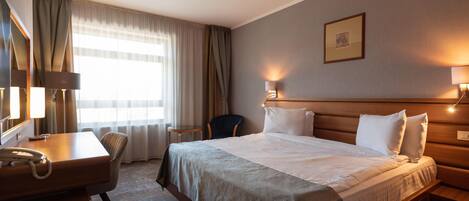 Habitación de hotel con cama, lámparas de noche, escritorio con teléfono, una silla y ventana con cortinas.