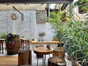 An outdoor seating area with wooden tables and stools, surrounded by potted plants and a stone wall.
