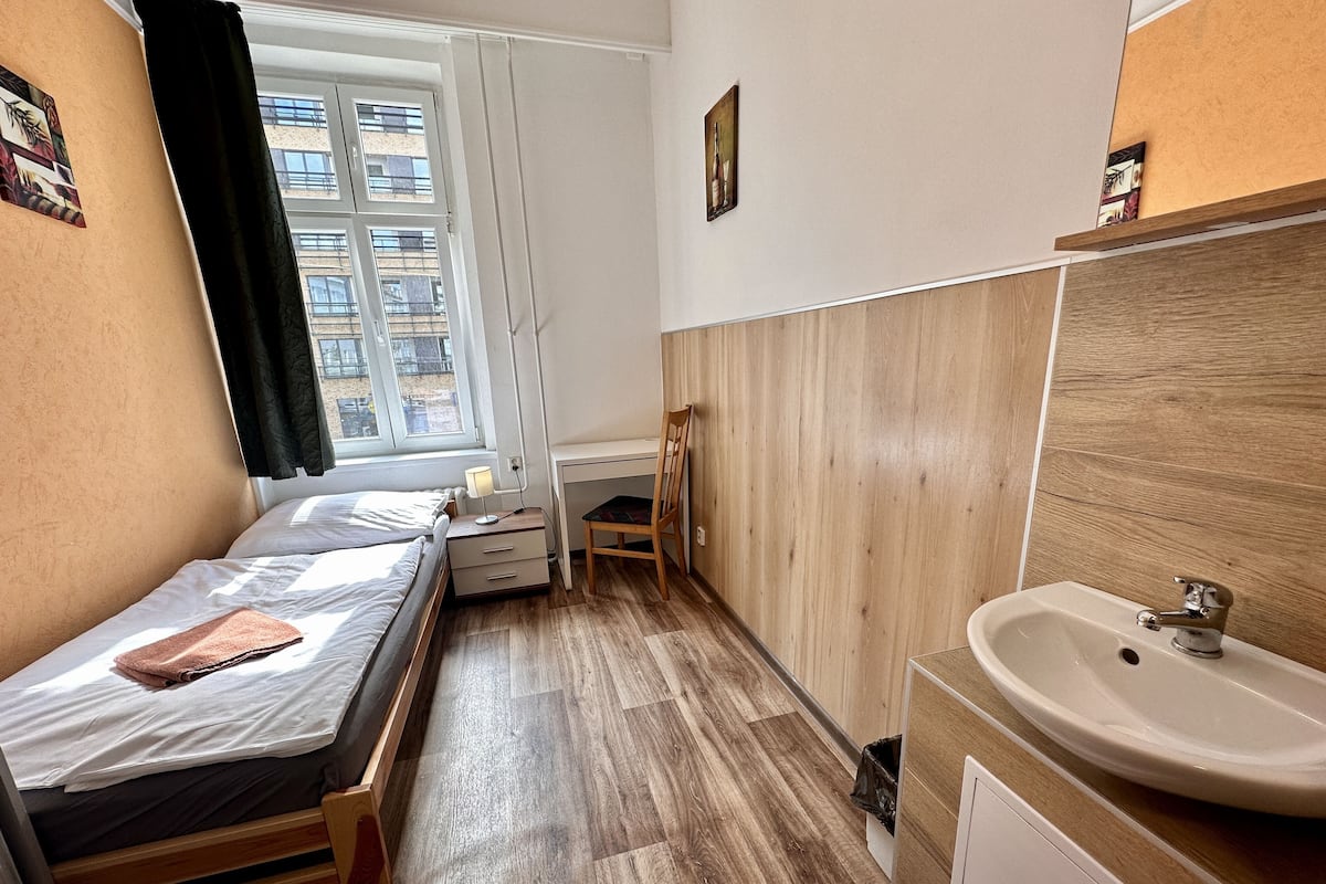A small, well-lit room with a bed, a desk, a chair, a window with curtains, and a sink.