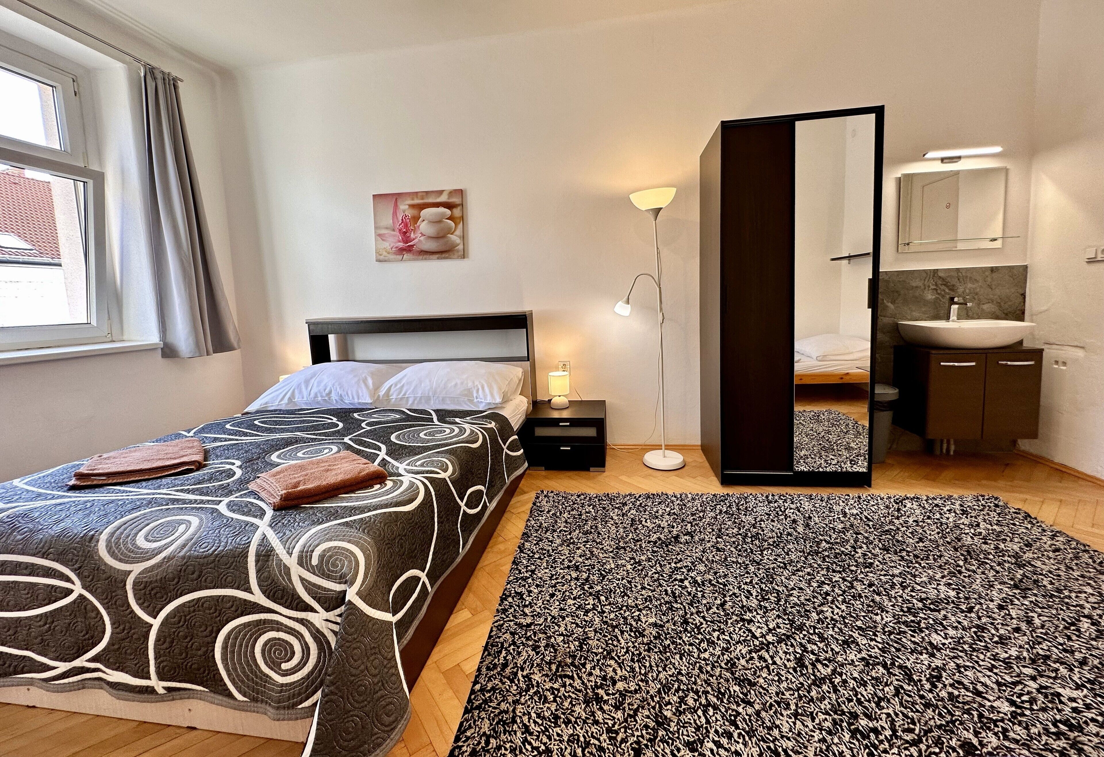 A bedroom with a bed, bedside table, lamp, and a bathroom mirror.