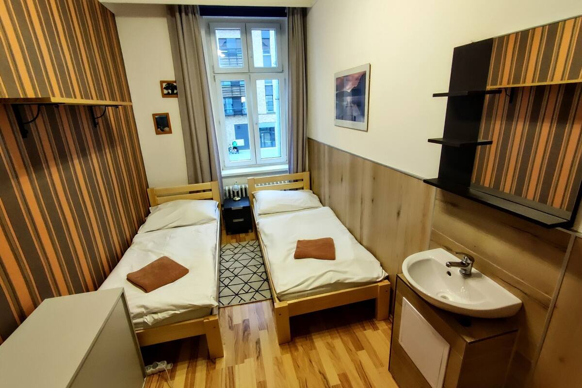 A hotel room with two wooden beds, a window with curtains, a small radiator, a sink, and a mirror.
