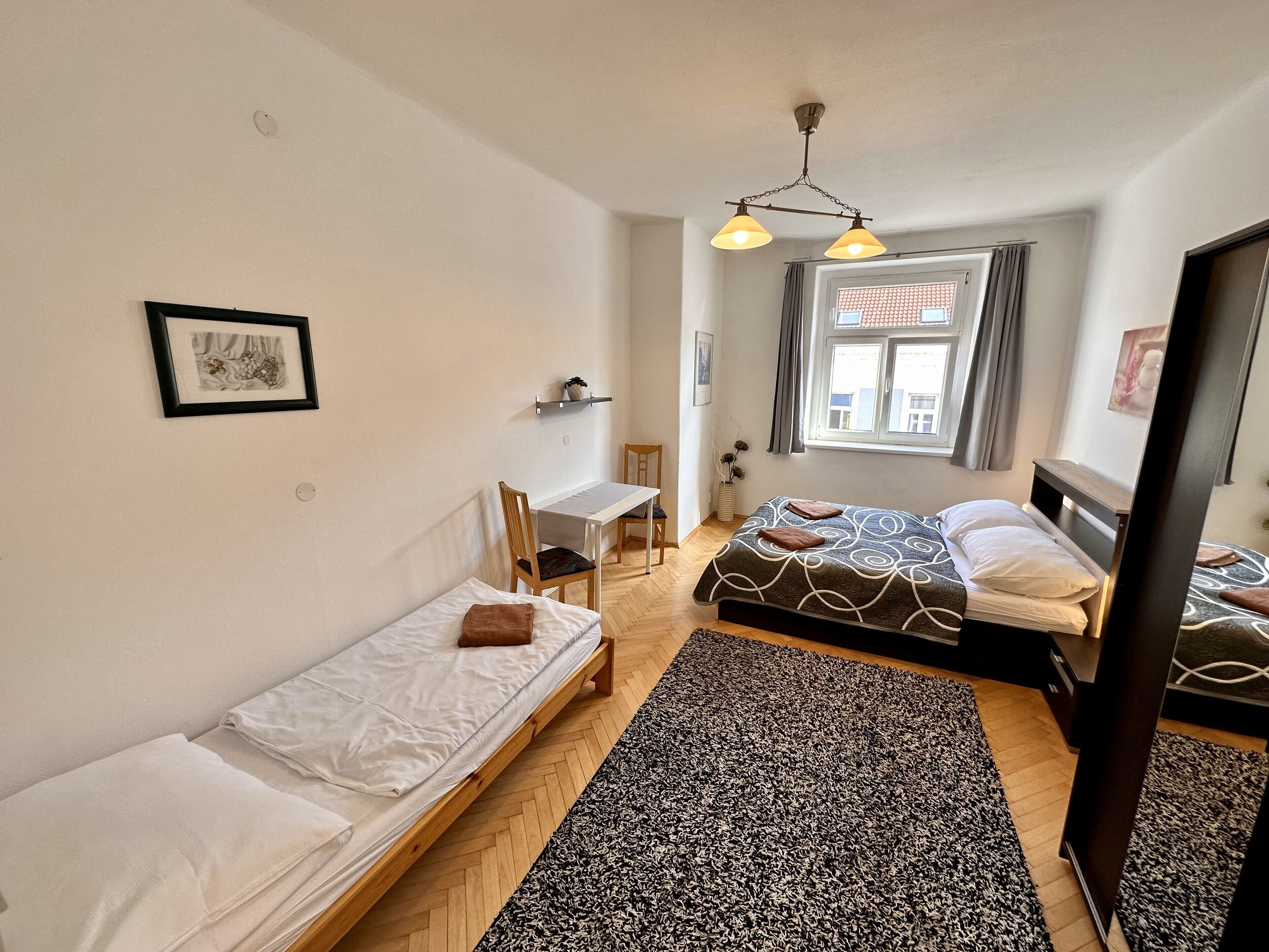 A small, well-lit room with two single beds, a desk, and a chair. There is a window with curtains, a framed picture on the wall, and a patterned rug on the floor.