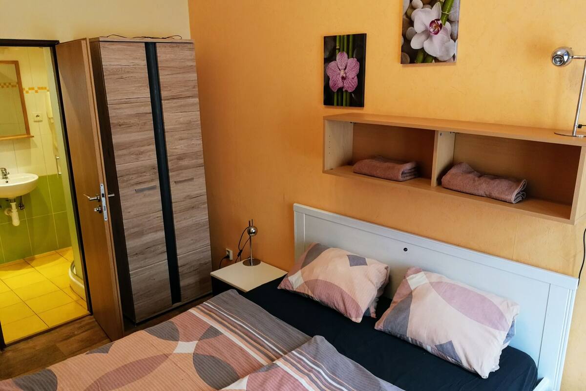 A bedroom with a bed, pillows, a nightstand, a mirror, a wardrobe, and a shelf with towels.