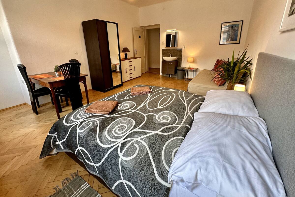 A bedroom with a bed, a desk, a wardrobe, a mirror, a sink, and a plant.