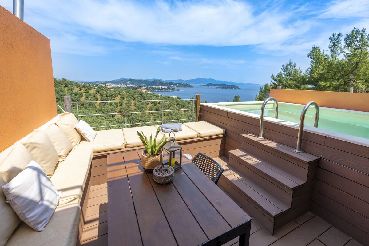 A balcony with a wooden table, a cushioned seating area, and a view of a body of water and greenery.