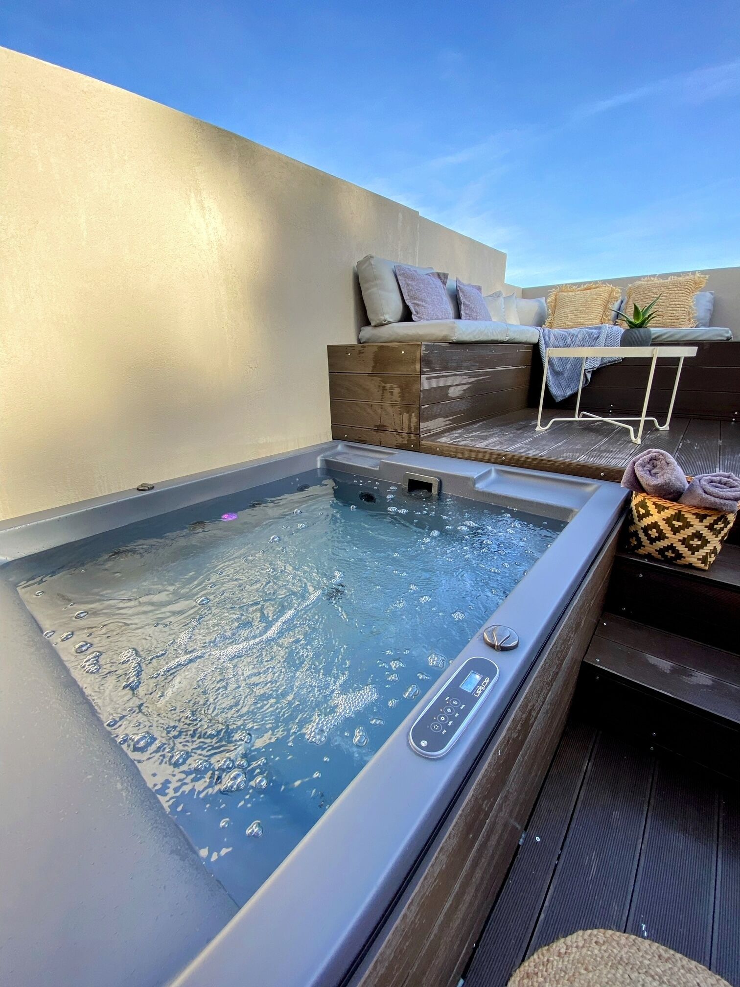 A hot tub with a control panel, wooden deck, and seating area with cushions.