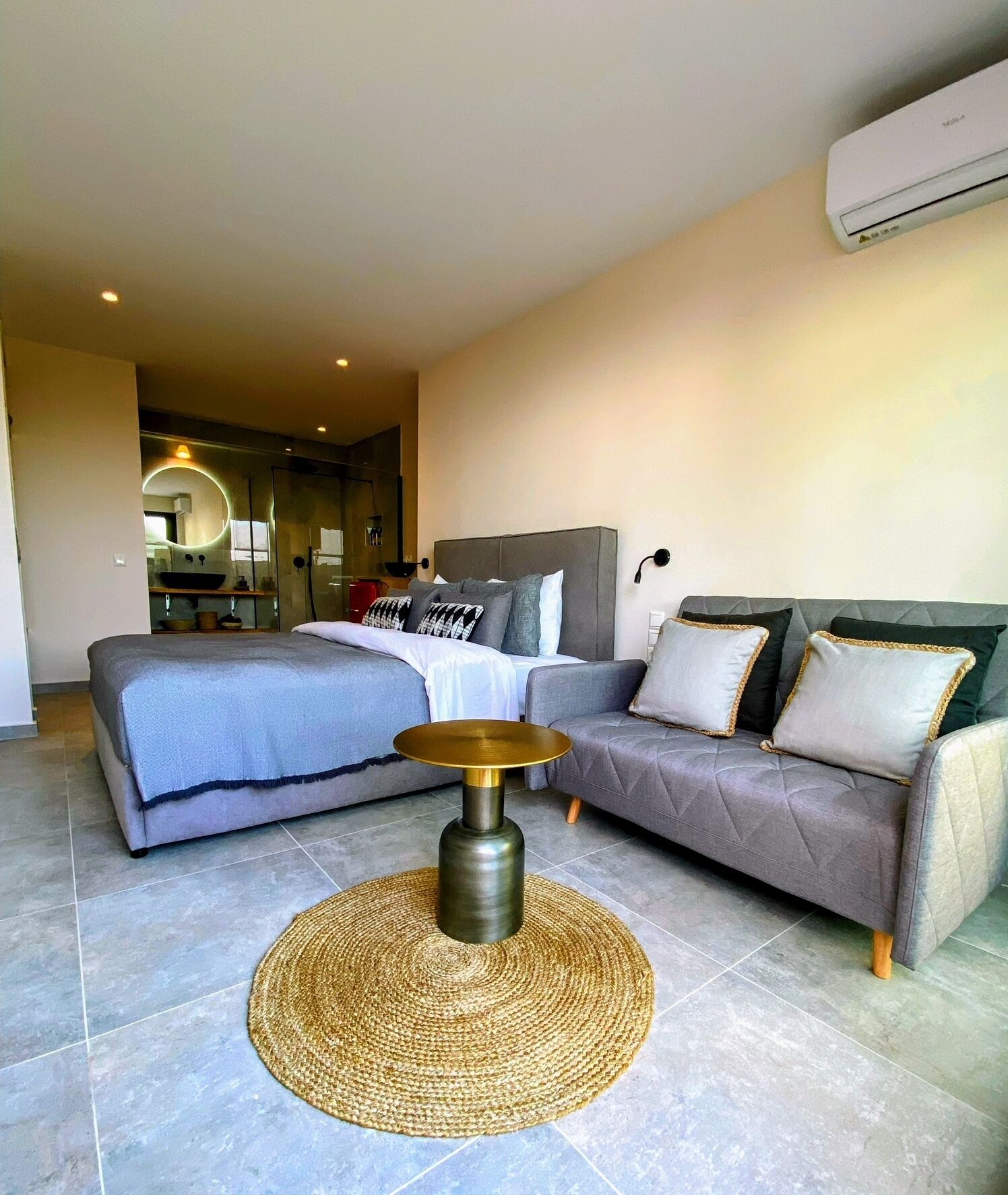 A modern bedroom with a large bed, a grey sofa, and a round woven table.