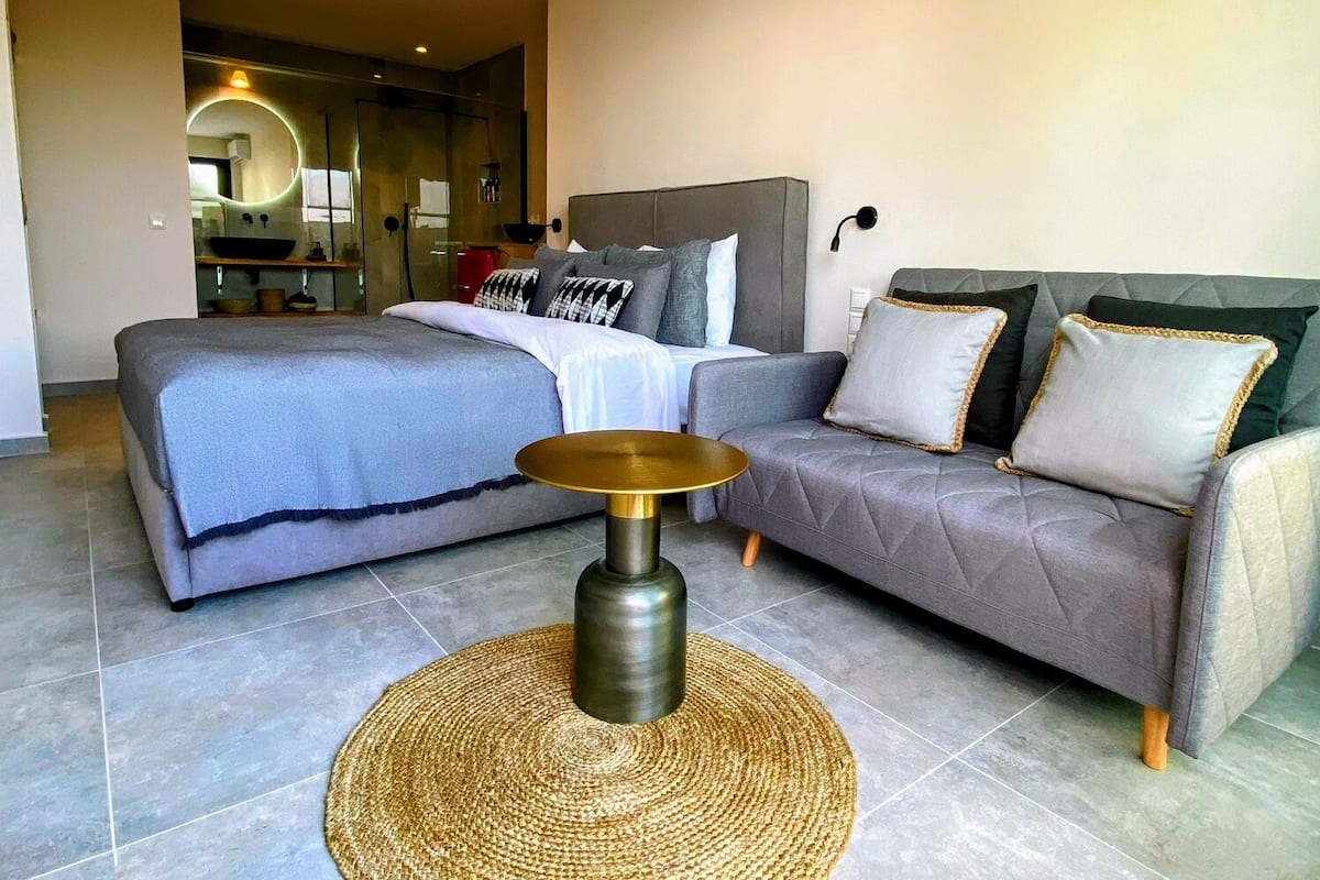 A modern bedroom with a large bed, a grey sofa, and a round woven table.