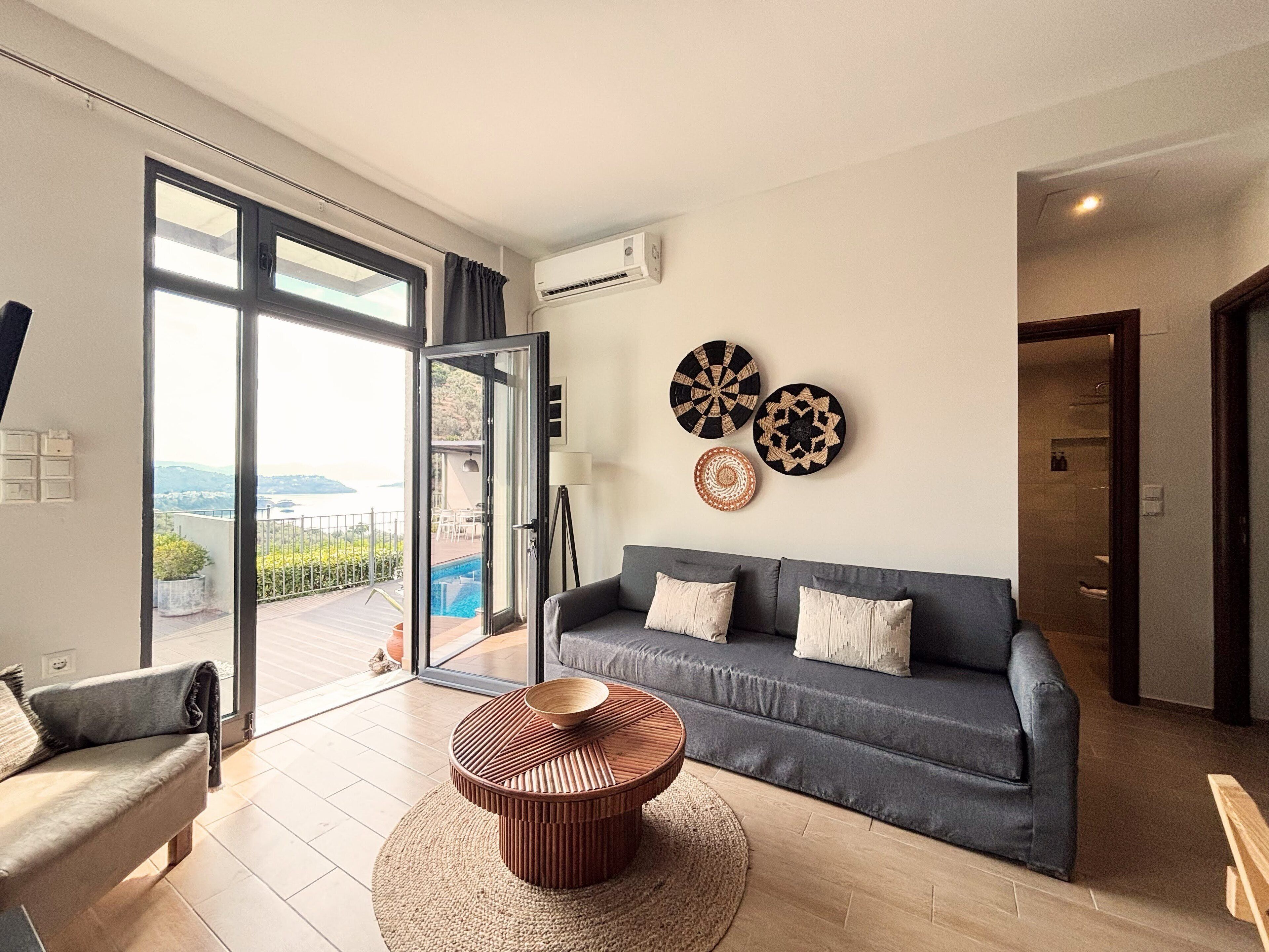 A modern living room with a grey sofa, a round wooden coffee table, and a view of a swimming pool through a glass door.