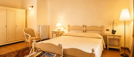 A bedroom with a wooden bed, a chair, a bedside table with a lamp, a radiator, and a wardrobe.