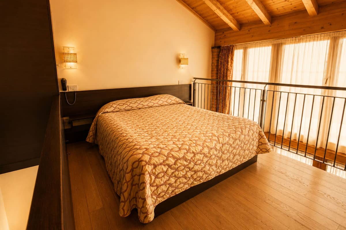 Une chambre avec un plafond en bois, un lit recouvert d’une couvre-lit à motifs, une table de chevet avec une lampe et une fenêtre avec des rideaux.