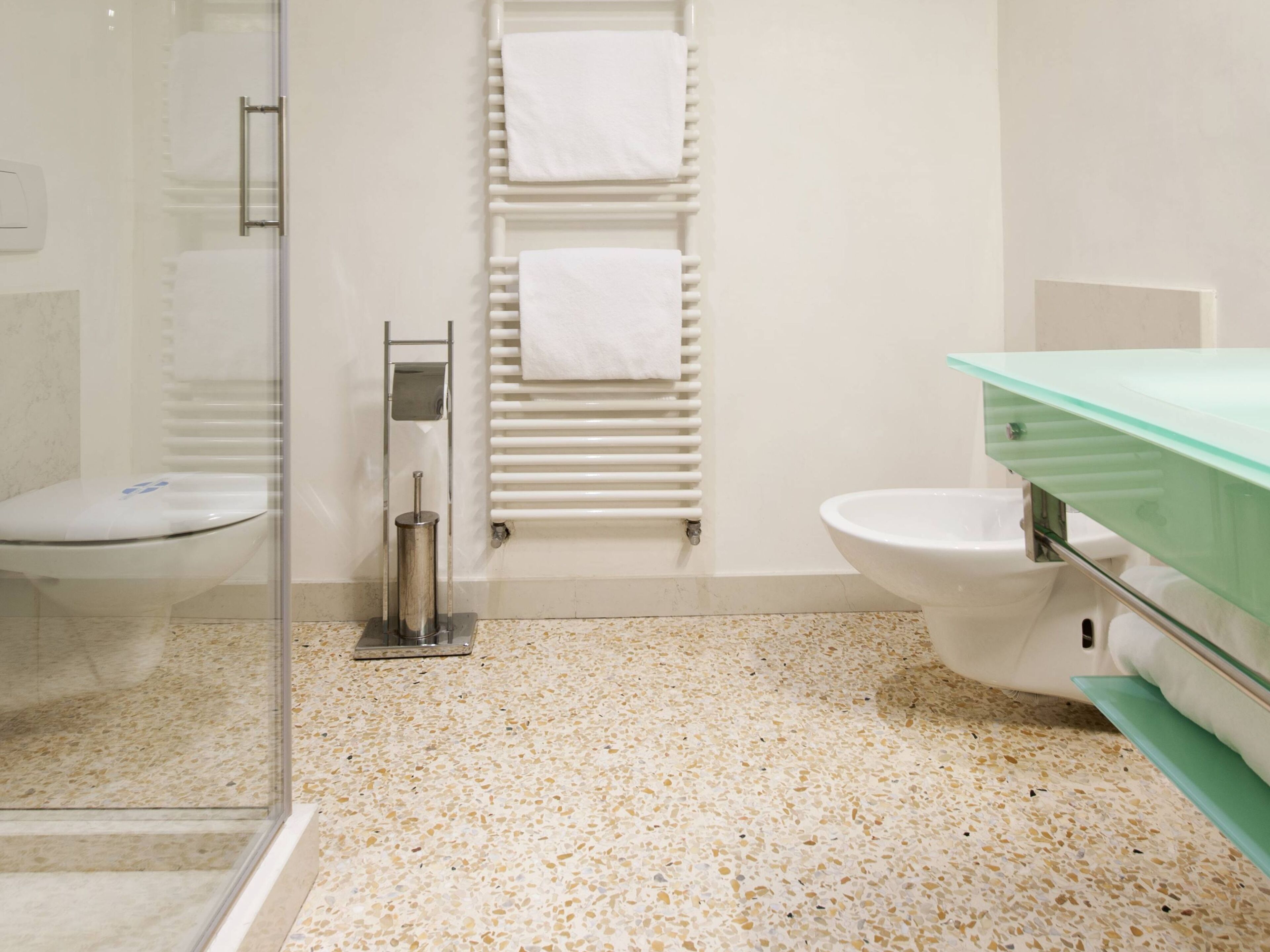 A bathroom with a glass shower enclosure, a toilet, a bidet, and a glass sink with a green countertop.