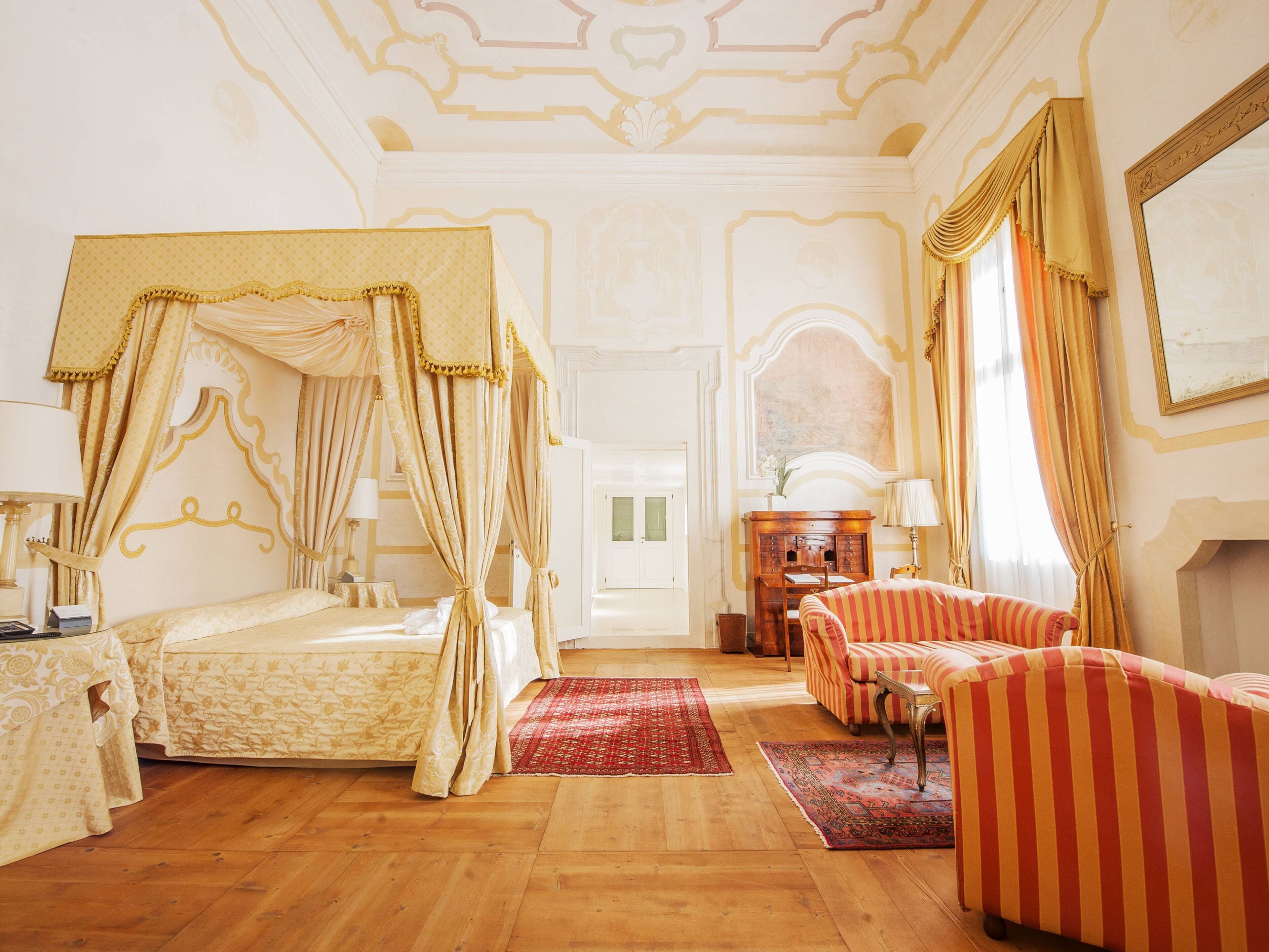 A four-poster bed with a canopy, a sofa, a small table, and a cabinet in a room with wooden flooring and ornate ceiling details.